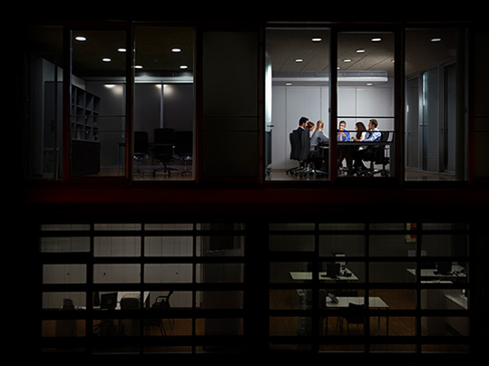 wide shot of a dark office with employees in a conference room working through the night